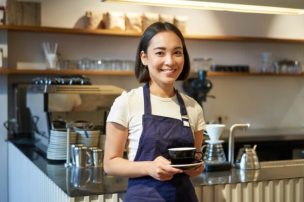 Portrait d'une femme asiatique souriante serveuse de café barista tenant une tasse de café servant des clients prenant