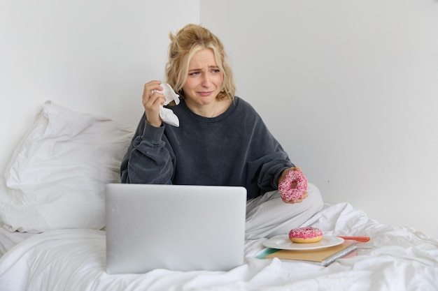 Photo gratuite portrait d'une femme assise sur un lit avec un ordinateur portable mangeant un donut et pleurant d'une scène de film triste