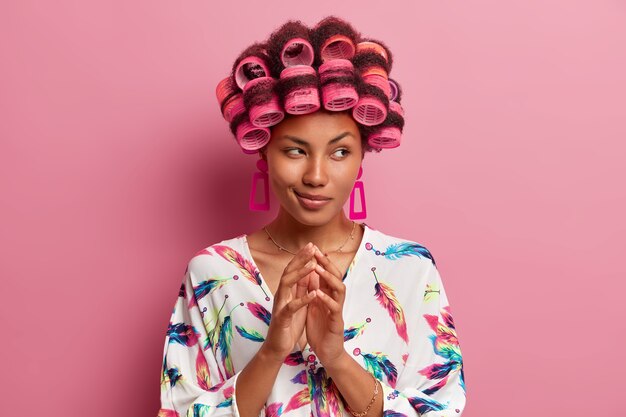 Photo gratuite portrait de femme au foyer réfléchie avec des rouleaux, des doigts croisés et des plans quelque chose, a l'air intrigué, porte une robe domestique