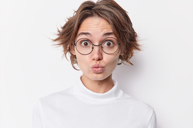 Portrait d'une femme aux cheveux noirs surprise qui boude les lèvres comme si elle voulait embrasser quelqu'un porte des lunettes rondes pour la correction de la vision cavalier décontracté isolé sur fond blanc. Expressions du visage humain