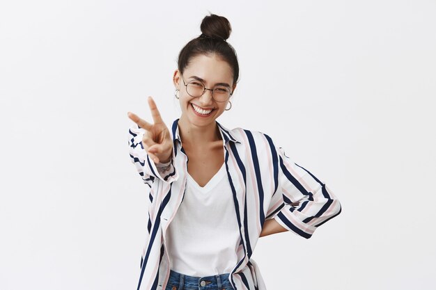 Photo gratuite portrait de femme belle et joyeuse optimiste à lunettes et chemise élégante, tirant la main avec le signe de la paix, souriant largement du bonheur et de l'amusement