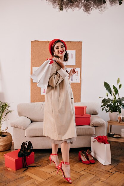 Portrait de femme en béret rouge et manteau beige. Jolie dame en vêtements d'automne légers et chapeau lumineux détient des achats dans l'appartement.