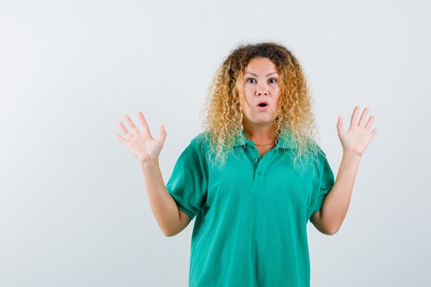 Portrait d'une femme blonde aux cheveux bouclés montrant un geste d'abandon en T-shirt vert et à la vue de face choquée