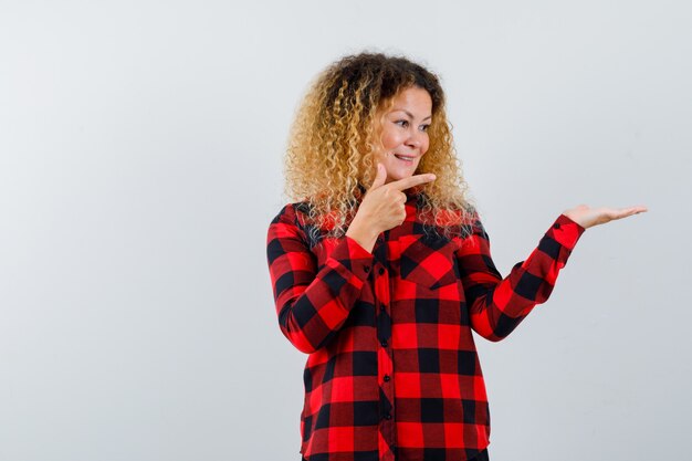 Portrait de femme blonde aux cheveux bouclés pointant sur sa paume écartée en chemise à carreaux et à la vue de face joyeuse