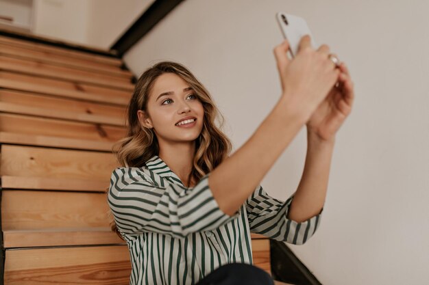 Portrait d'une femme blonde bouclée en chemise de soie rayée blanche et verte sourit et prend le selfie à la maison