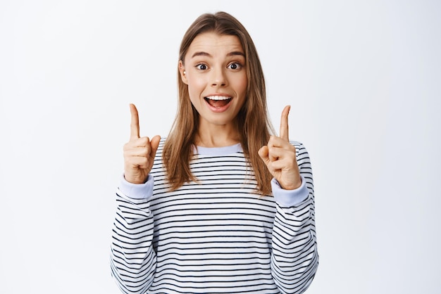 Portrait d'une femme blonde excitée en chemise rayée pointant les doigts vers le haut et vous parlant de la publicité regardant la caméra avec un sourire heureux fond blanc