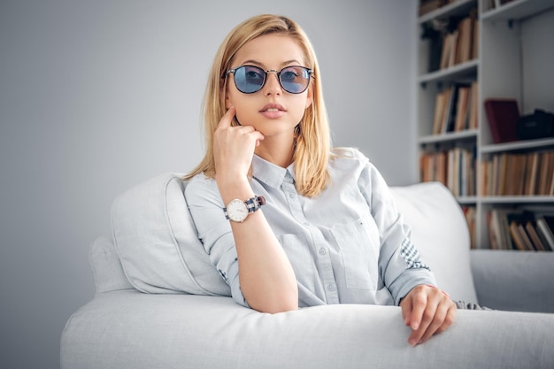 Portrait d'une femme blonde en lunettes de soleil bleues hipster est assise sur un canapé dans une pièce avec des stands de livres.