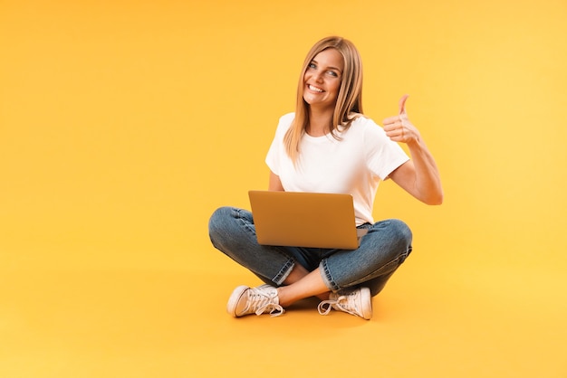 Portrait d'une femme blonde montrant le pouce vers le haut et utilisant un ordinateur portable assis avec les jambes croisées isolées sur un mur jaune en studio