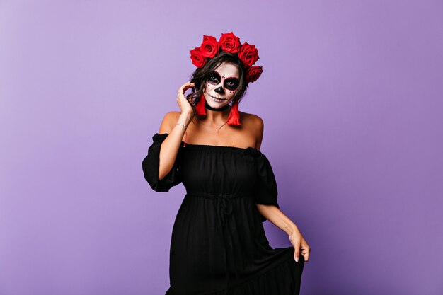 Portrait de femme bronzée latine gracieuse au look Halloween. Fille en robe noire touche ses boucles d'oreilles rouge vif