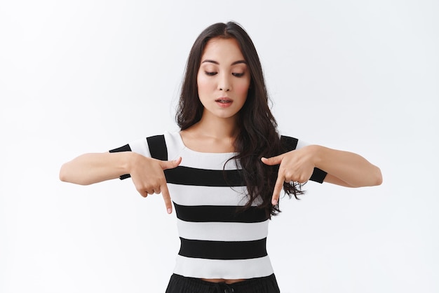 Portrait d'une femme brune asiatique intense et interrogée en t-shirt rayé retenant son souffle regardant et pointant vers le bas la bouche ouverte regardant attentivement et curieux fond blanc