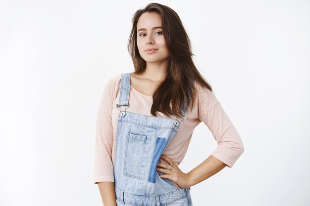 Portrait d'une femme brune impertinente et élégante avec de longs cheveux naturels en salopette en jean posant, tenant la main sur la taille et souriante confiante et sûre d'elle à l'avant sur un mur gris.