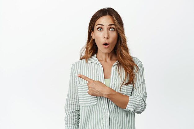 Portrait d'une femme brune impressionnée en chemisier, gasp dire WOW, pointant le doigt vers la gauche à la bannière promotionnelle de vente, choquée par une grande remise, fond blanc