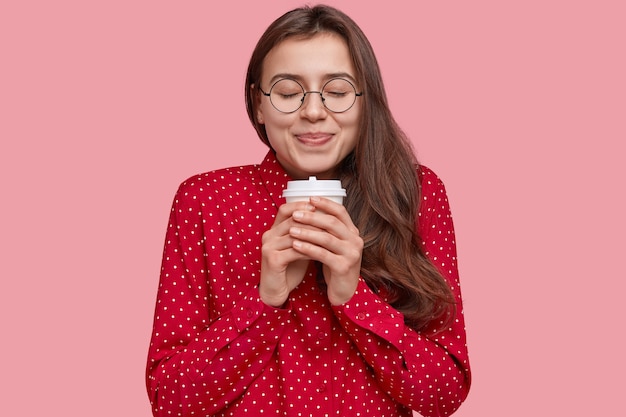 Portrait de femme brune joyeuse aux yeux fermés, tient le café à emporter, réchauffe les mains, a une expression romantique