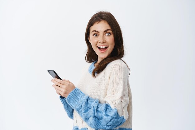 Portrait d'une femme brune joyeuse tourne la tête à la caméra après avoir lu les nouvelles sur l'écran du téléphone portable souriant excité à l'aide de l'application smartphone fond blanc