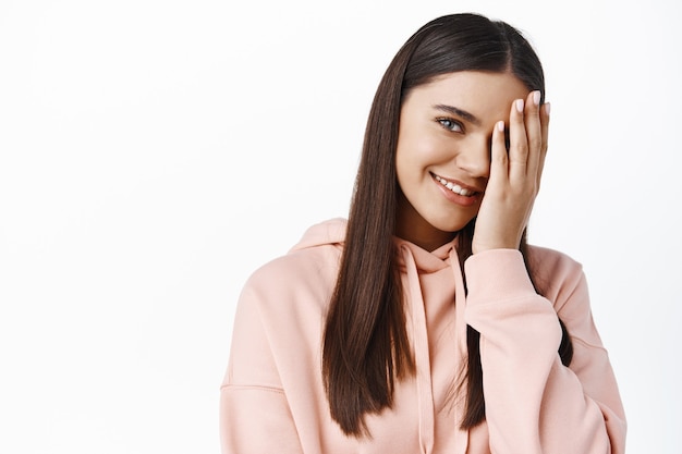 Portrait d'une femme brune souriante couvrant la moitié du visage et semblant heureuse, portant du maquillage à la lumière naturelle, mur blanc