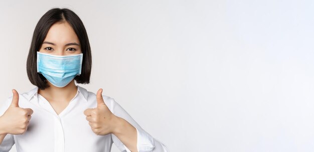 Portrait d'une femme de bureau asiatique souriante dans un masque médical montrant les pouces vers le haut recommandant qch debout sur fond blanc