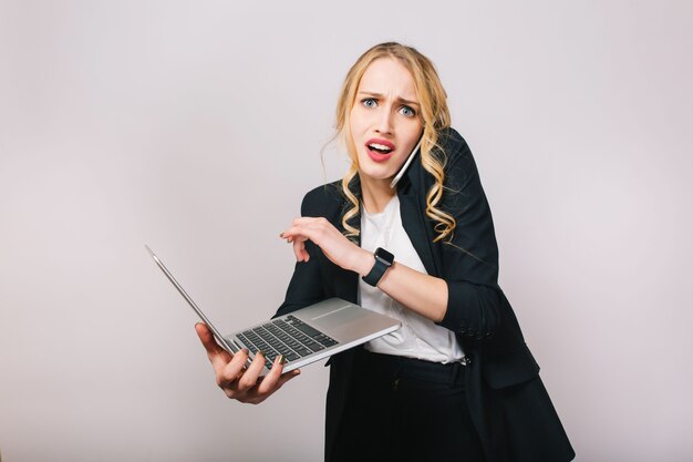 Portrait femme de bureau jolie blonde moderne en chemise blanche et veste noire travaillant avec ordinateur portable, parler au téléphone. Étonné, être en retard, bouleversé, se rencontrer, exprimer de vraies émotions