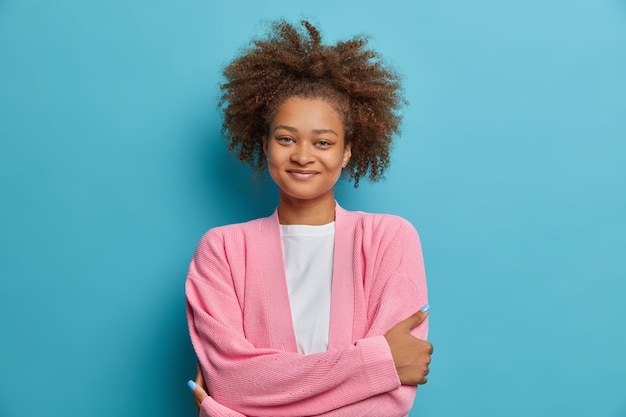 Photo gratuite portrait de femme calme et agréable avec une expression satisfaite garde les bras croisés a les cheveux bouclés touffus peignés dans la queue écoute agréablement un ami a une conversation décontractée avec quelqu'un.