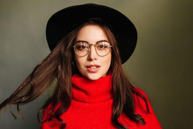 Portrait de femme caucasienne aux cheveux noirs en pull rouge et chapeau à larges bords sur mur gris.