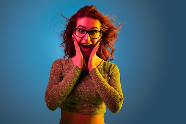 Portrait de femme caucasienne isolé sur fond bleu studio en néon. Beau modèle féminin aux cheveux rouges dans un style décontracté. Concept d'émotions humaines, expression faciale, ventes, publicité. Étonné.