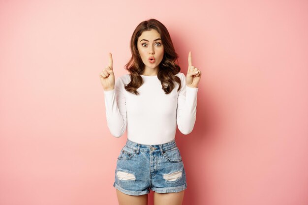 Portrait d'une femme caucasienne surprise, fille élégante pointant les doigts vers le haut et semblant impressionnée, montrant le logo du magasin, publicité à prix réduit, fond rose