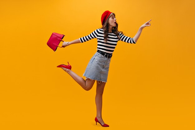 Portrait de femme en chemise rayée posant étonnamment sur fond orange. Fille cool en béret rouge et jupe tendance s'amuse.