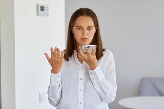 Portrait d'une femme confuse et perplexe portant une chemise blanche posant à la maison avec un téléphone intelligent dans les mains, levant le bras, ne comprend pas pourquoi l'appareil ne fonctionne pas.