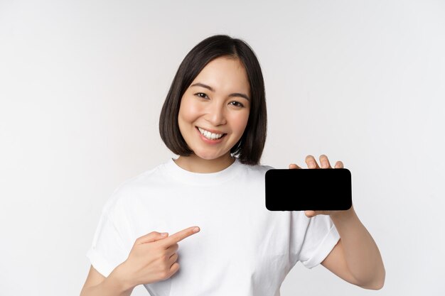 Portrait d'une femme coréenne souriante pointant le doigt sur l'écran du téléphone portable montrant l'affichage horizontal du smartphone recommandant le site Web ou le magasin en ligne fond blanc