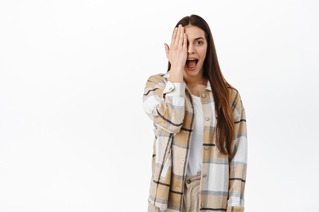 Portrait d'une femme couvrant la moitié de son visage et ayant l'air surpris par un autre, haletant impressionné et excité, vérifiant l'offre promotionnelle impressionnante, voir l'offre impressionnante dans la boutique, mur blanc