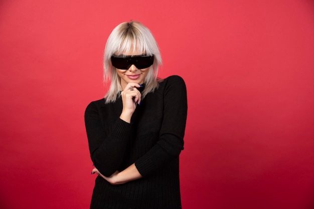 Portrait de femme debout et posant avec des lunettes noires sur un mur rouge.