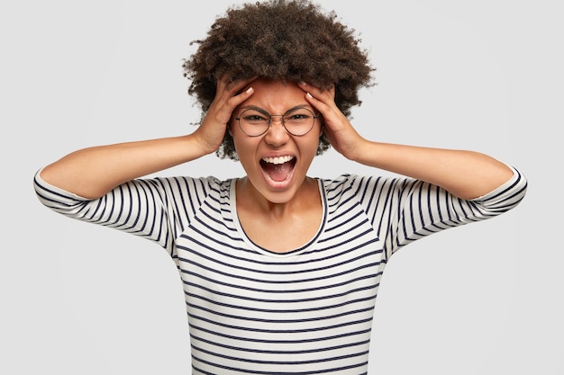 Portrait de femme déprimée à la peau sombre mécontente avec coupe de cheveux afro, s'exclame avec des sentiments négatifs, garde les mains sur les tempes, se sent désespérée, vêtue d'une veste rayée, isolée sur un mur blanc