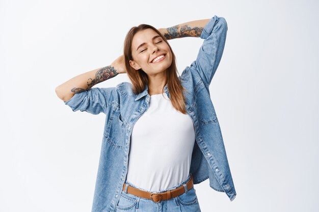 Portrait d'une femme détendue et insouciante aux cheveux blonds, tenant les mains derrière la tête et les yeux fermés avec un sourire heureux, se reposant satisfait, debout contre un mur blanc