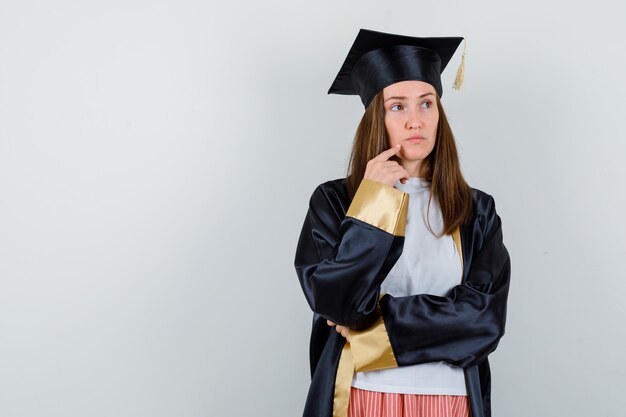 Portrait de femme diplômée tenant le doigt sur le menton dans des vêtements décontractés, uniforme et à la vue de face réfléchie