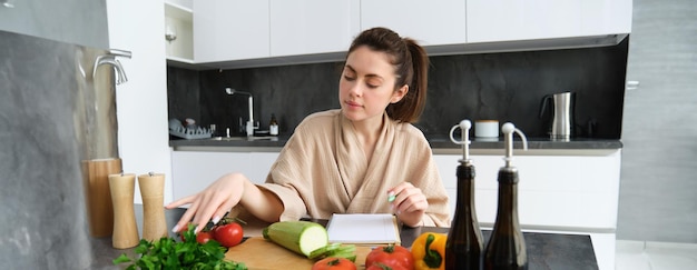 Photo gratuite portrait d'une femme écrivant une liste de produits d'épicerie prenant des notes dans une recette assise dans la cuisine à proximité
