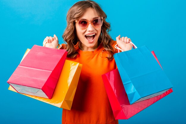 Portrait de femme élégante souriante séduisante excitée accro du shopping en robe à la mode orange tenant des sacs sur fond bleu studio isolé