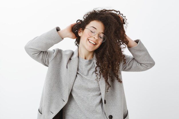 Portrait de femme émotive heureuse insouciante en manteau gris et lunettes, touchant et agitant les cheveux bouclés mignons, souriant joyeusement les yeux fermés