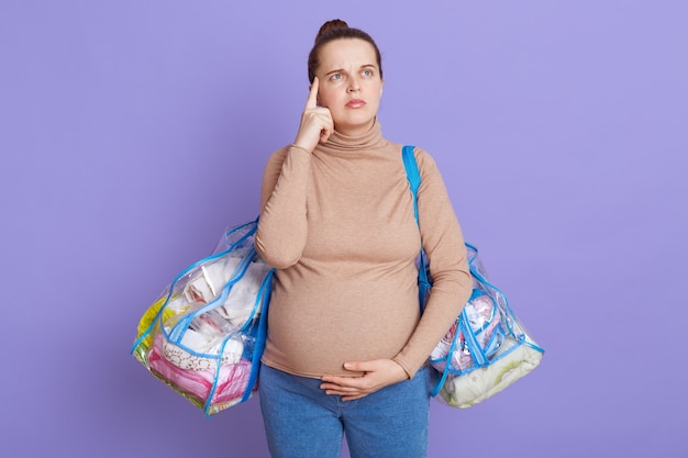 Portrait de femme enceinte réfléchie, essayant de se souvenir qu'elle emmène tout à l'hôpital, garde le doigt sur la tempe, levant les yeux avec une expression pensive, isolée sur un mur lilas