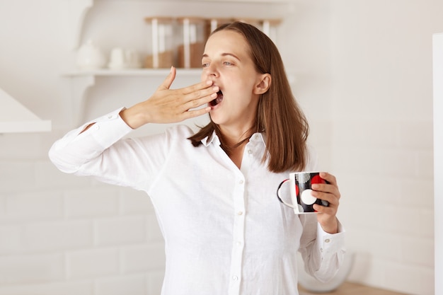 Portrait d'une femme endormie debout dans la cuisine et regardant ailleurs, bâillant, couvrant la bouche avec la main, se réveillant tôt et buvant du thé ou du café chaud dans une tasse.