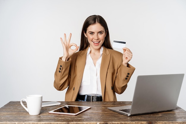 Portrait d'une femme entrepreneur assise à une table de bureau, montrant une carte de crédit et un pouce levé, recommandant, debout sur fond blanc.