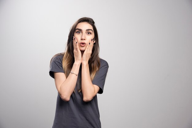 Portrait de femme étonnée touchant son visage posant sur fond gris.