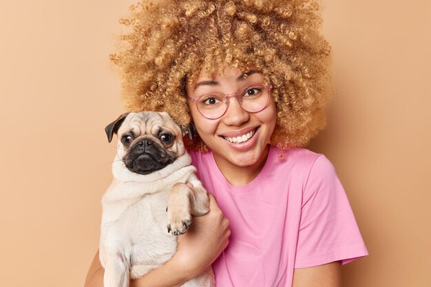 Portrait d'une femme européenne heureuse aux cheveux bouclés blonds pose avec un chien carlin heureux que le propriétaire de l'animal pose pour faire une photo mémorable vêtue d'un t-shirt décontracté rose isolé sur fond beige