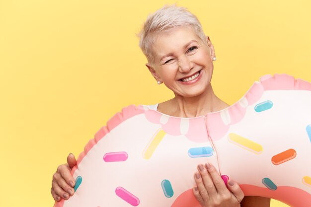 Portrait de femme européenne mature joyeuse émotionnelle avec une coiffure de lutin transportant un cercle rose gonflable en forme de beignet, faisant une grimace drôle comme si elle prenait une morsure, s'amusait, profitait de l'heure d'été