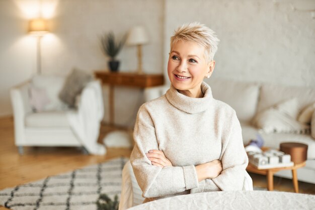 Portrait de femme européenne mature magnifique avec une coiffure courte se détendre à la maison assis à table dans le salon en croisant les bras sur la poitrine en essayant de se réchauffer dans un pull à col roulé en cachemire confortable, souriant