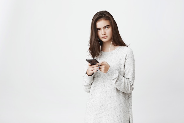 Portrait de femme européenne mignonne inquiète et frustrée, tenant un smartphone et portant des écouteurs