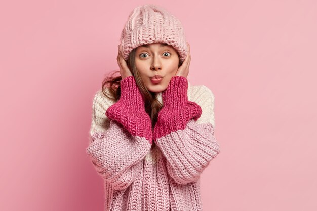 Portrait de femme européenne porte un couvre-chef tricoté, vêtu d'un pull ample, veut embrasser quelqu'un, isolé sur un mur rose. Style décontracté. Concept de mode d'hiver. Expressions faciales humaines