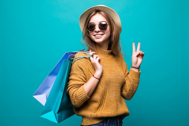 Portrait d'une femme excitée heureuse dans des vêtements colorés lumineux tenant des sacs à provisions en position debout et montrant le geste de paix isolé sur mur vert