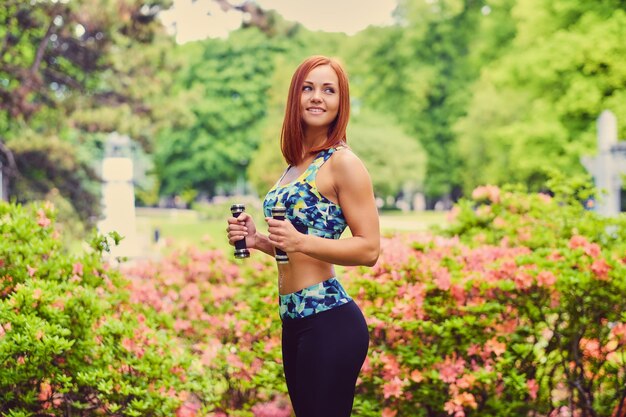 Portrait de femme fitness rousse détient des haltères dans un parc extérieur.