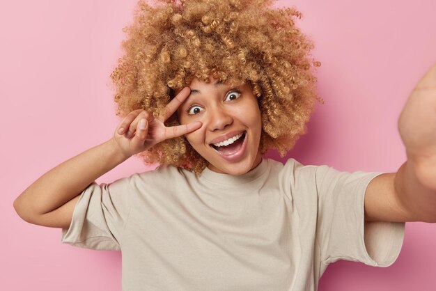 Portrait d'une femme gaie aux cheveux bouclés et touffus prend selfie et fait des gestes de paix à la caméra sourit positivement vêtue d'un t-shirt décontracté isolé sur fond rose Une femme ludique capture une photo