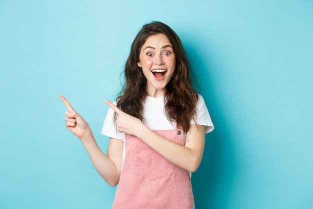 Portrait d'une femme heureuse et excitée avec des boucles sombres et brillantes, souriante fascinée, pointant les doigts vers la bannière du logo, montre une publicité, debout sur fond bleu.