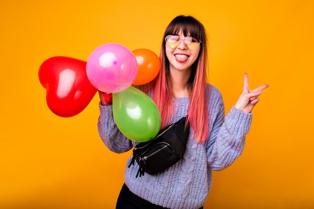 Portrait de femme heureuse jeune hipster montrant le geste ok et rire, pull bleu confortable, lunettes à la mode et sac, tenant des ballons colorés, ambiance de fête.